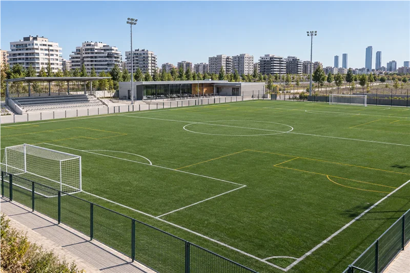Campo de fútbol de césped artificial con graderío pequeño y edificios residenciales al fondo.