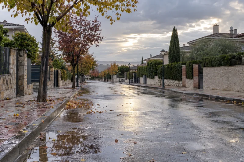 Calle residencial mojada después de una lluvia intensa, con charcos sobre la calzada.
