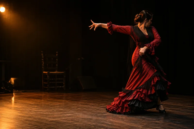 Bailaora con vestido rojo y negro extendiendo un brazo sobre un escenario de madera iluminado en tonos ámbar.