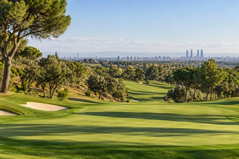 Campo de golf con una calle verde, árboles y el perfil urbano de Madrid al fondo.