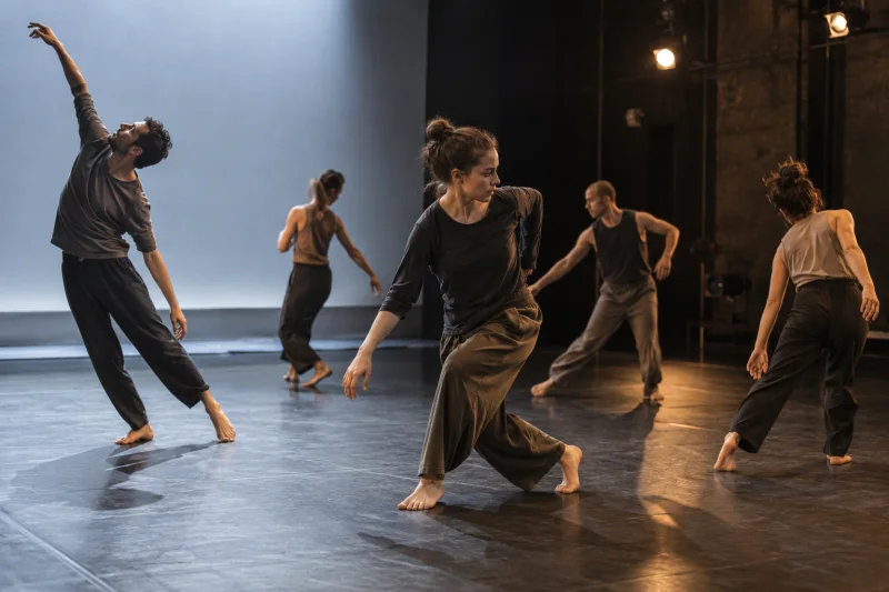 Cinco intérpretes ensayan danza contemporánea en un escenario de caja negra con luz fría a un lado y luz cálida al otro.