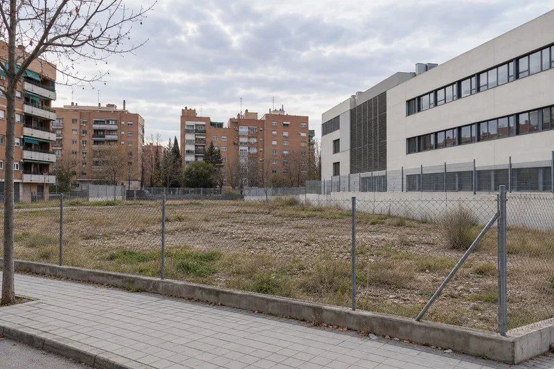 Solar urbano vacío tras una valla metálica, junto a bloques residenciales y un edificio institucional.